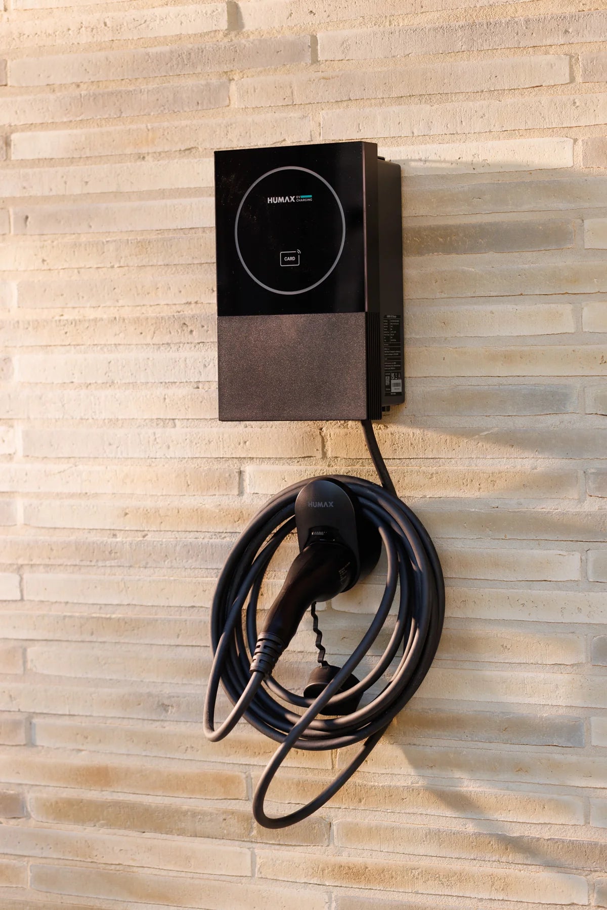 Humax MX7 7kW tethered home EV charger in black wall-mounted on horizontal-tile stone wall with full Type 2 cable looped on Humax holster below