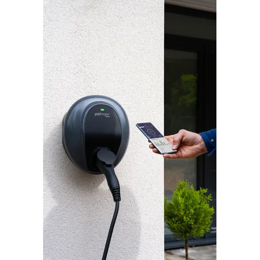 Pod Point Solo 3S EV charger mounted on exterior wall with person checking charging stats on smartphone app - UK home charging point