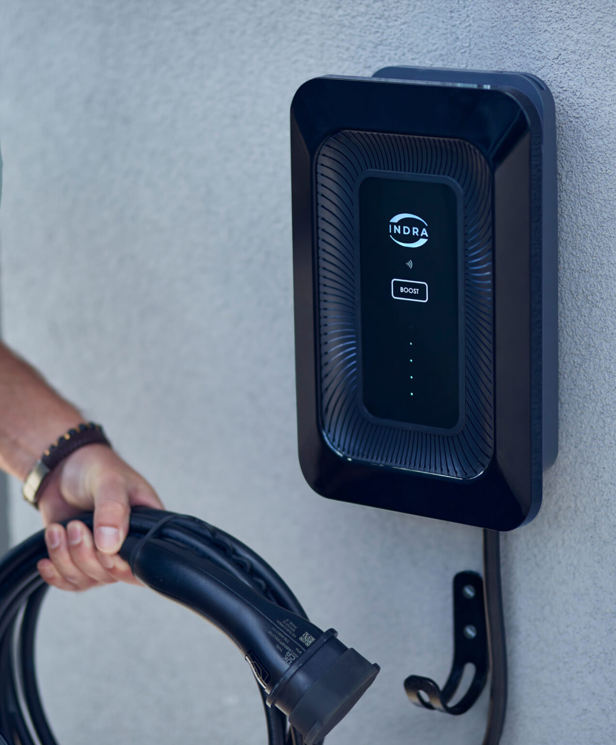 Indra Smart LUX home EV charger in white wall-mounted with sunburst face texture, INDRA logo, BOOST button and hand holding Type 2 connector lifestyle install shot