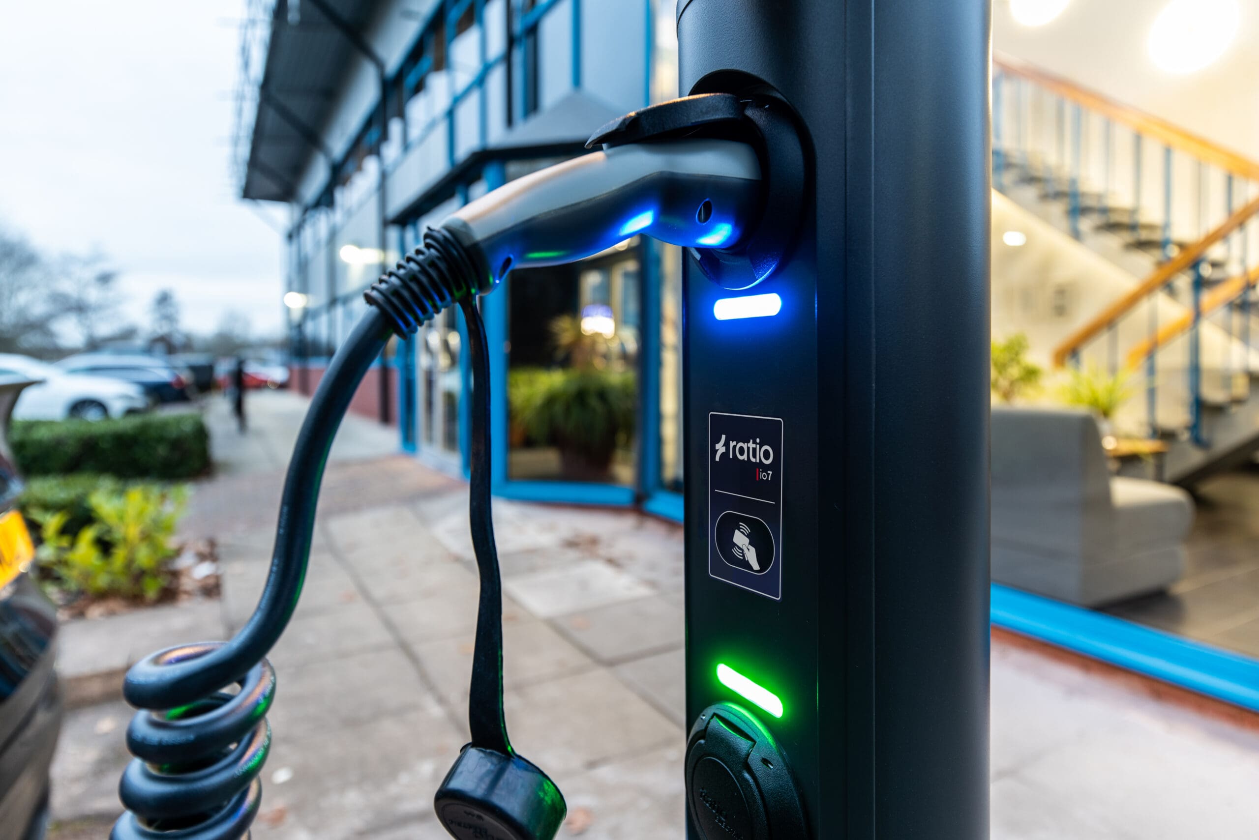 Ratio iO7 commercial EV charging bollard close-up with Type 2 cable in holder, illuminated blue and green LED indicators, ratio io7 branding and RFID logo