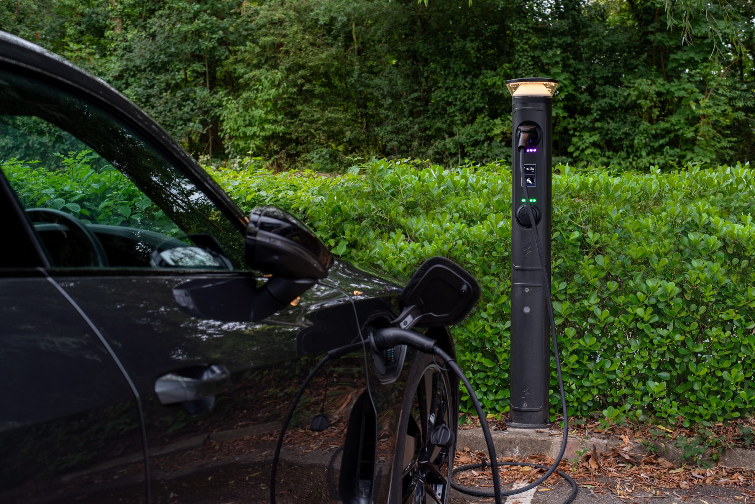 Ratio iO8 bollard EV charger installed beside hedge with EV plugged in via Type 2 cable, illuminated top cap visible against trees