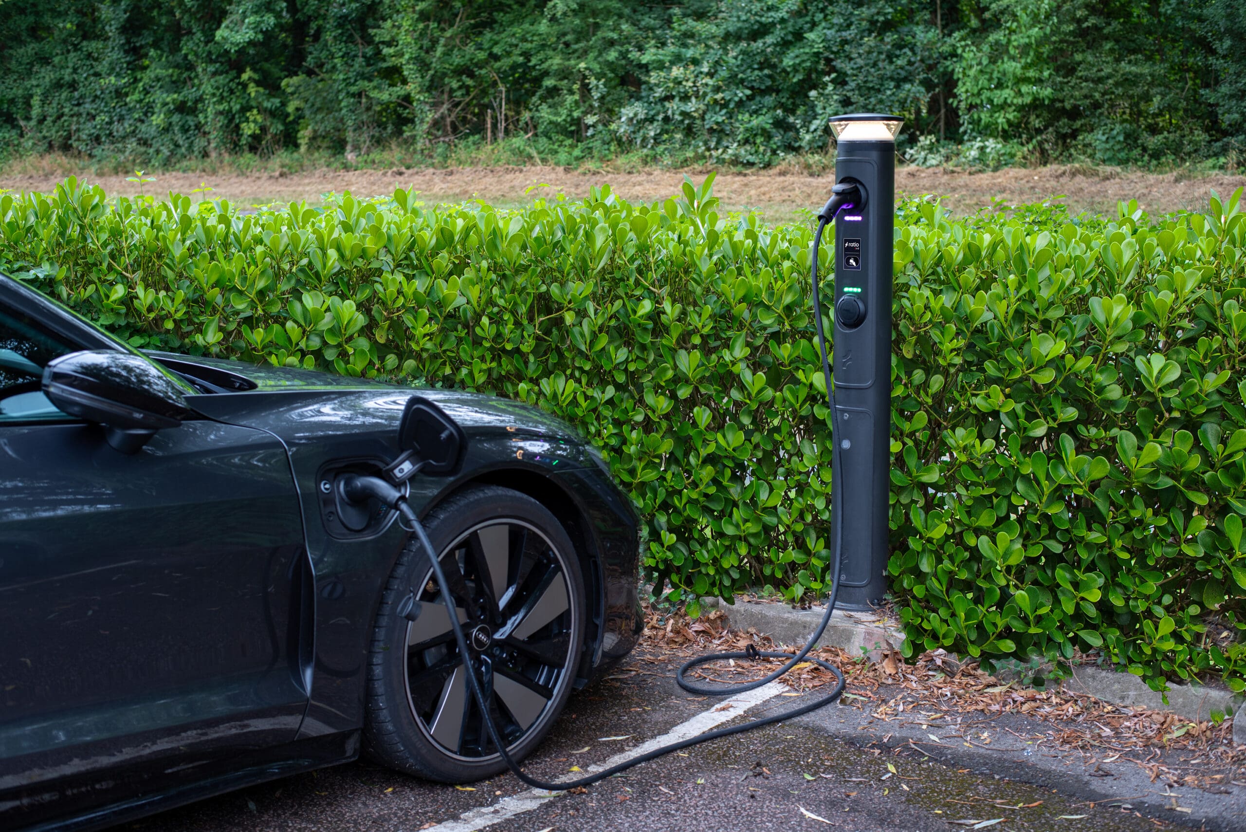 Ratio iO8 bollard EV charger with golden-glow illuminated top cap charging Porsche Taycan via Type 2 cable, hedge-lined kerbside install