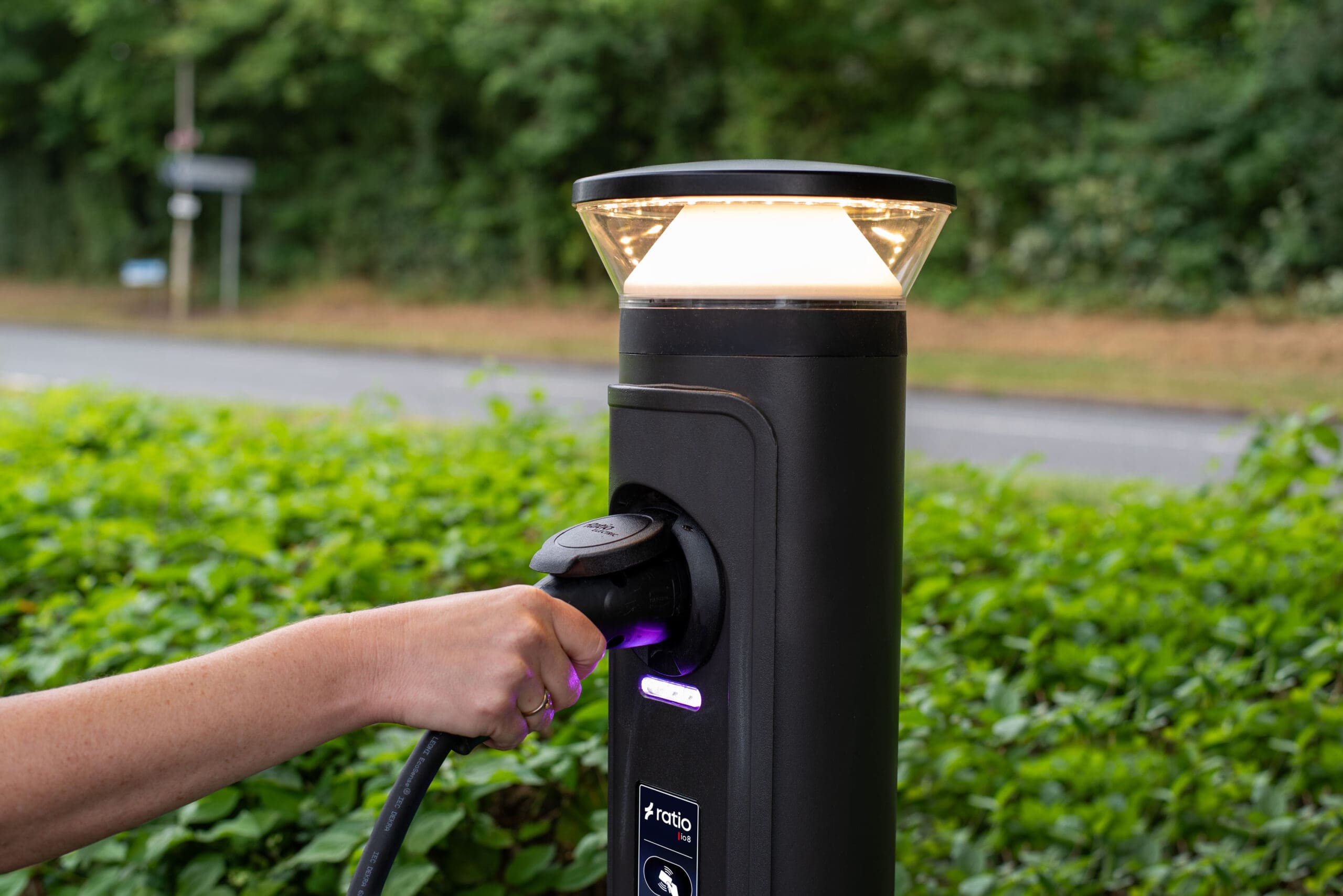 Person plugging Type 2 connector into Ratio iO8 bollard EV charger, illuminated golden top cap and purple LED at socket, outdoor commercial install